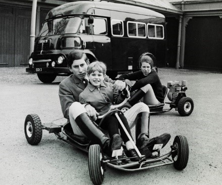 Photograph showing a young Prince Edward sharing a go-kart with the Prince of Wales who is controlling its pedals. Princess Anne is in middle ground on another go-kart. A small bus, with registration plate 'EGH 3', is in the background, parked in front of some garages.