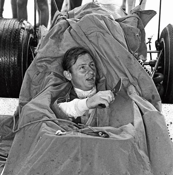 A well-armed Bruce McLaren emerges from a tarpaulin covering the M7 Formula One car during a rainy testing session at Brands Hatch in 1968.