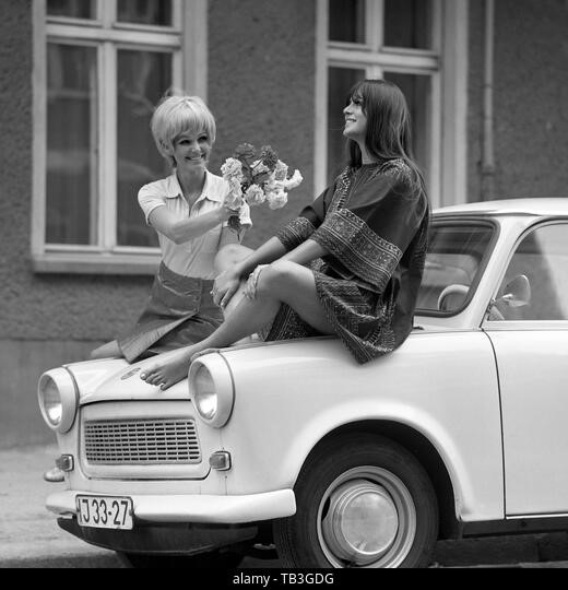 July 07, 1971, Berlin, GDR. A young woman hands flowers to her friend sitting on a Trabant. 