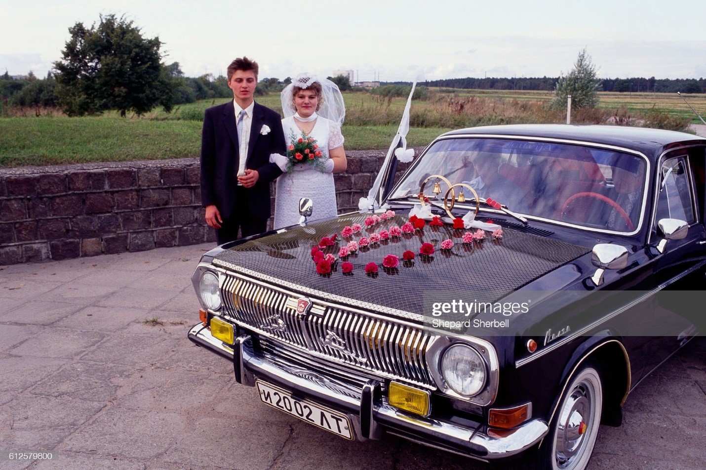 A bride and groom, in Latvia, pose for a portrait next to their bridal limousine decorated with flowers and large wedding rings in 1991. 