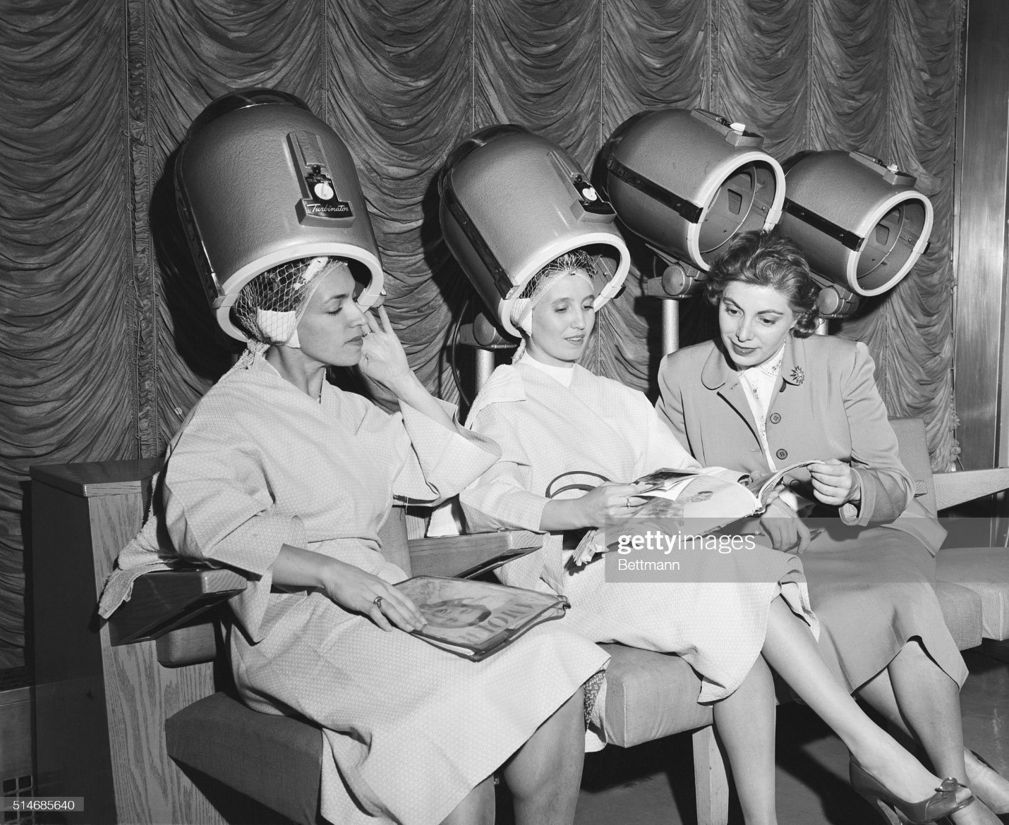 International News Service Women's Editor Olga Curtis explains an American fashion magazine to two Russian dancers as they sit under pink dryers at Lilly Dache's Salon in New York.