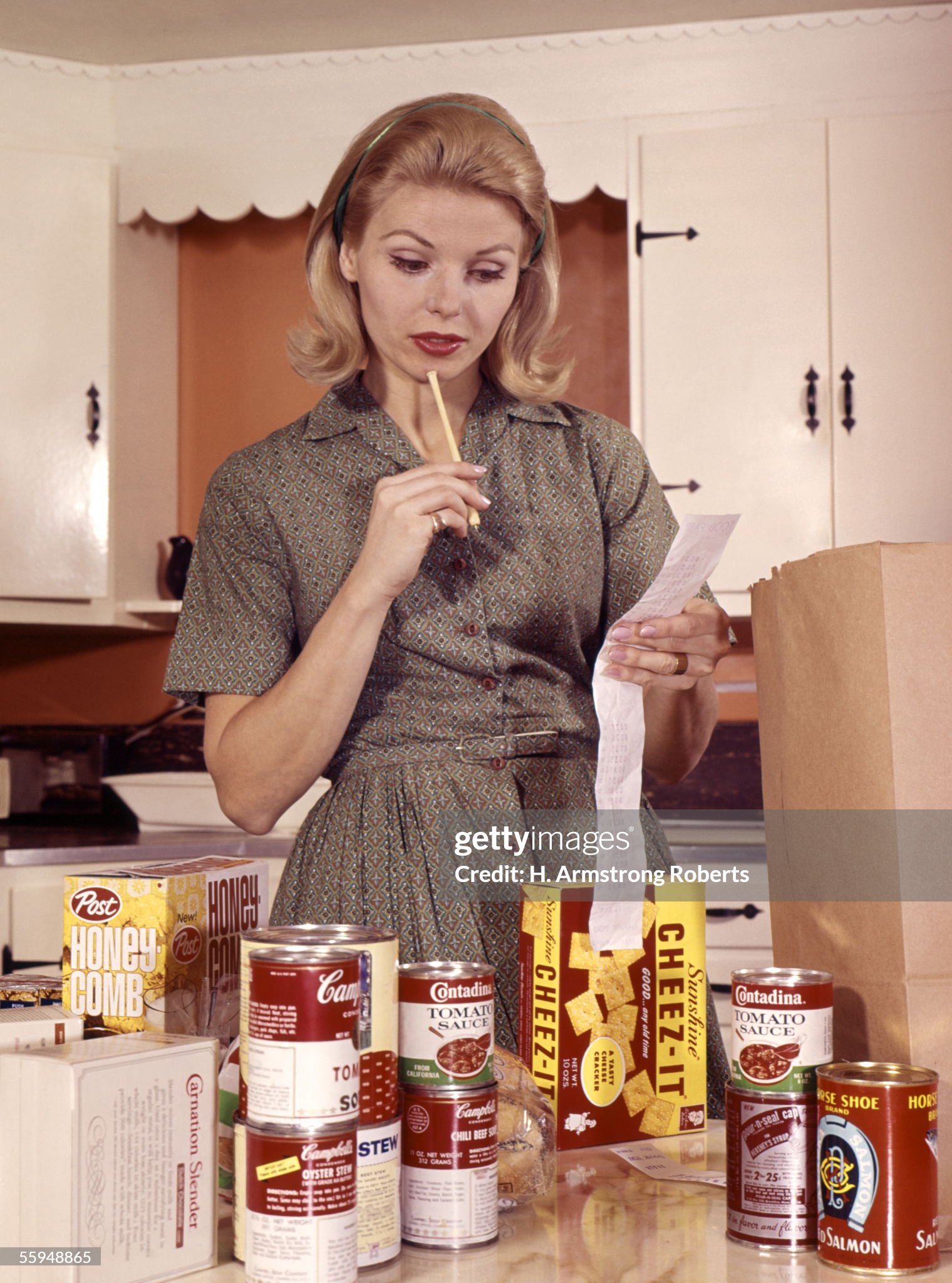 United States, circa 1960s. Blonde woman housewife in kitchen compare cash register receipt to items groceries on counter brown shopping bag. 