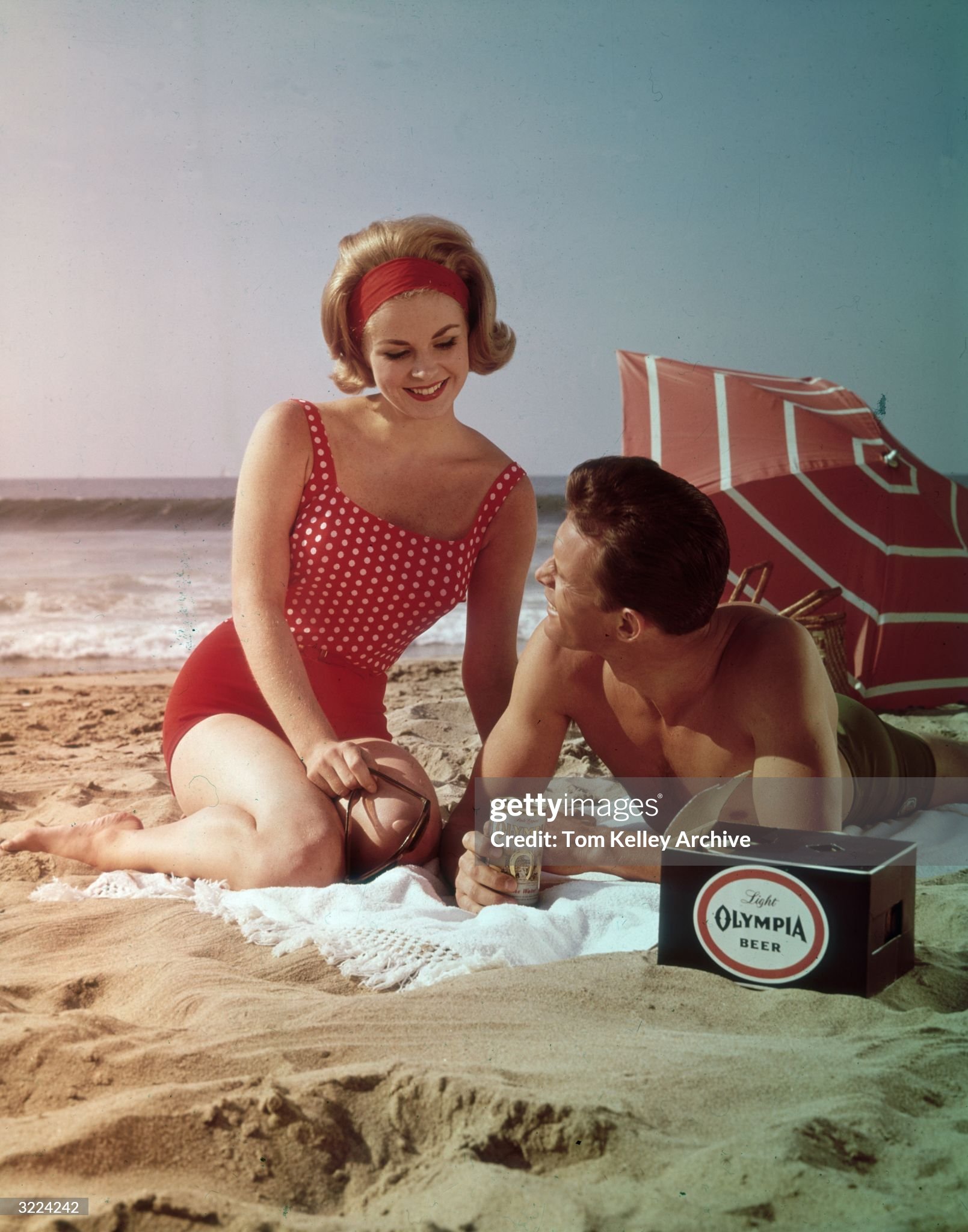 Circa 1960. A young couple in bathing suits pose next to a case of beer on a beach in an advertisement for Light Olympia Beer. 