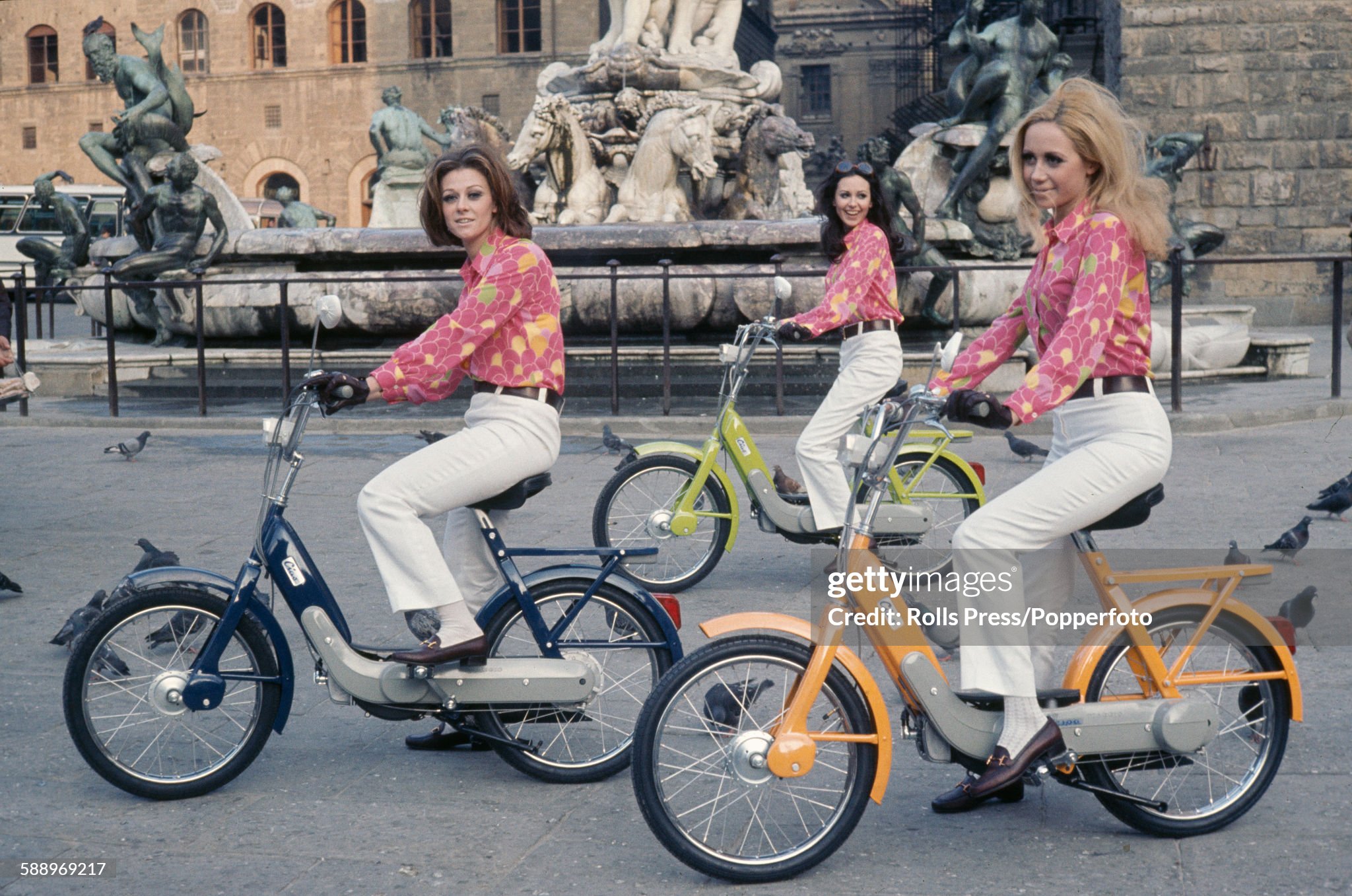 View of three female models wearing identical pink blouses and white trousers, pictured sitting on recently launched Piaggio Ciao mopeds in front of the Fountain of Neptune on the Piazza della Signoria in Florence, Italy, on 13th March 1968. 