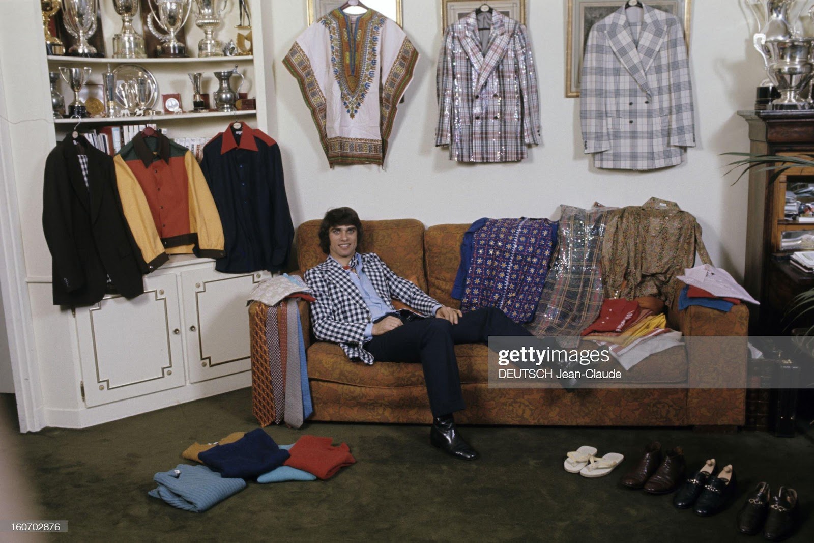 Men's Fashion, France, Paris, January 04, 1973. François Cevert poses half-lying down on a sofa, dressed in a black and white checkered jacket over a sky blue shirt with a scarf tied around his neck, over black trousers and leather boots, in a room with as many clothes and suits on hangers as there are trophies.