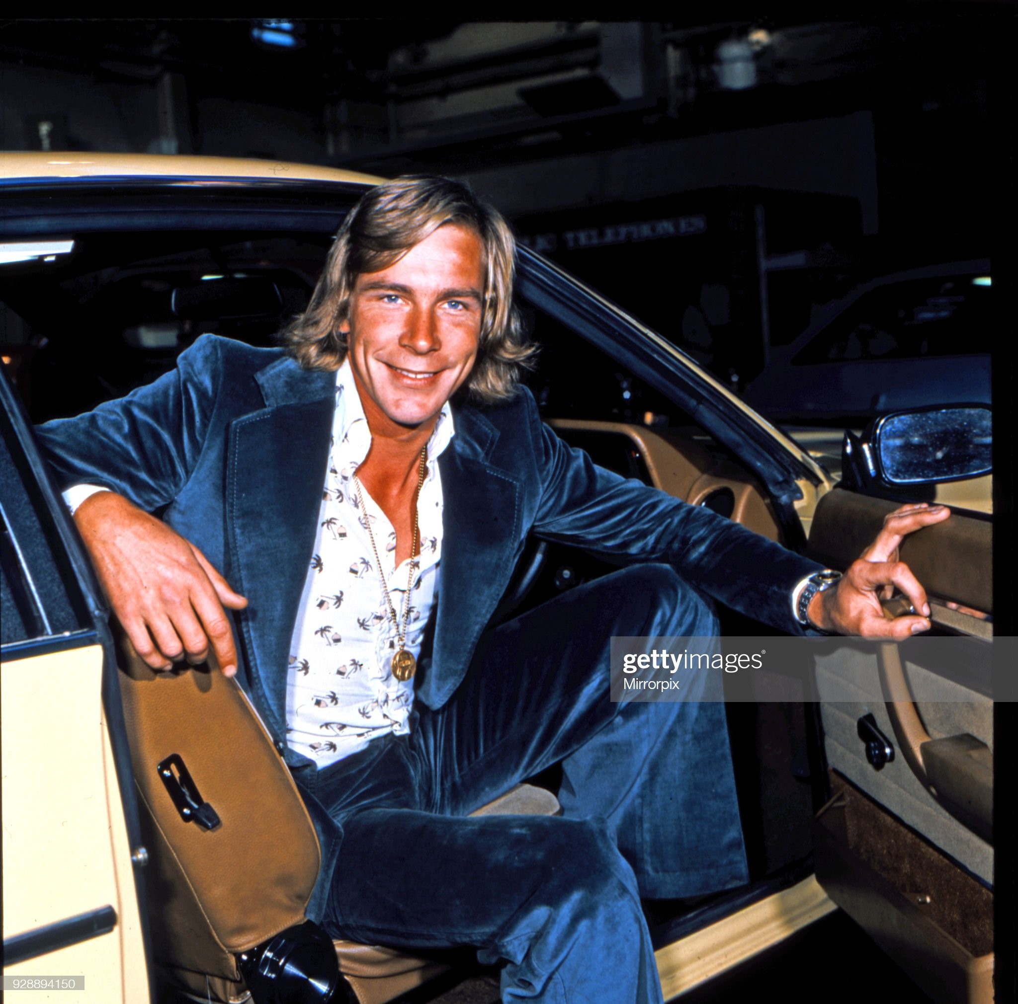 James Hunt, smiles to camera, whilst sitting in the front seat of his car in May 1978. James is wearing a medallion, white patterned shirt and blue velvet suit.
