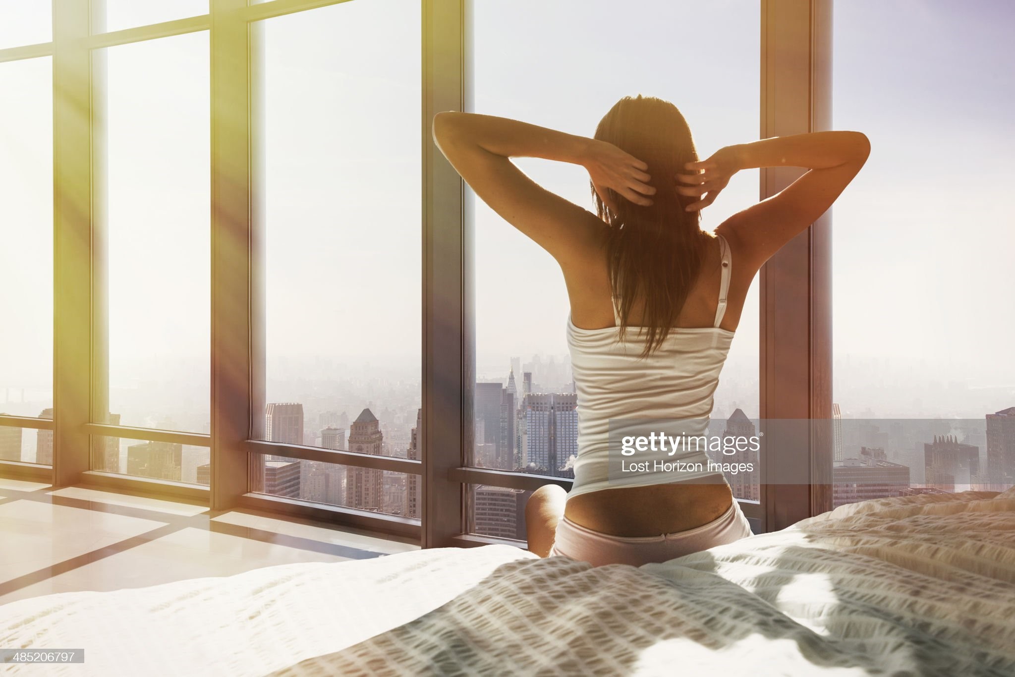 Young woman sitting on bed, stretching, overlooking city. 