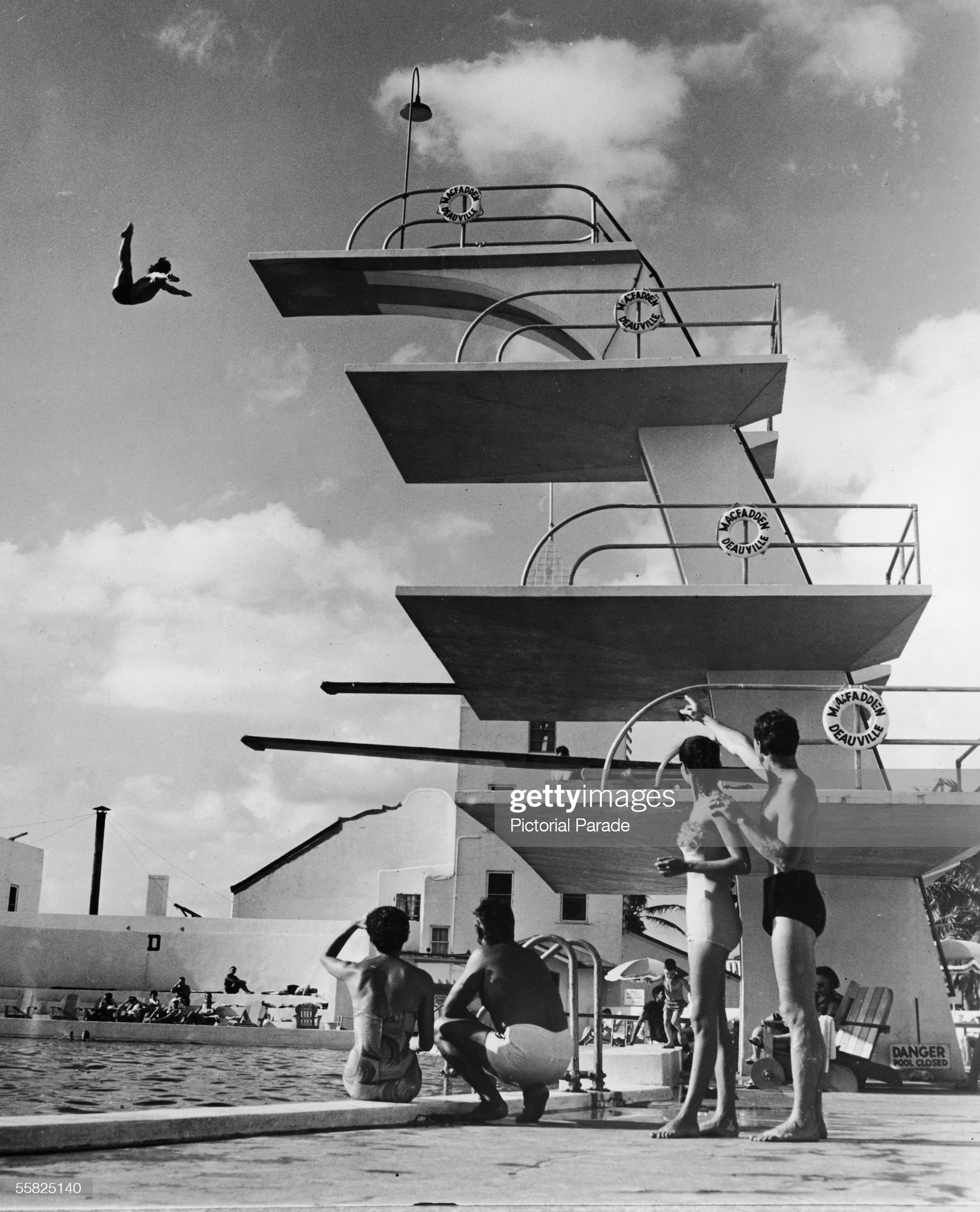 People sit poolside and watch as a swimmer dives off the high platform of the Olympic-size swimming pool at the MacFadden - Deauville Hotel (now the Deauville Beach Resort), Miami Beach, Florida, 1950s. 