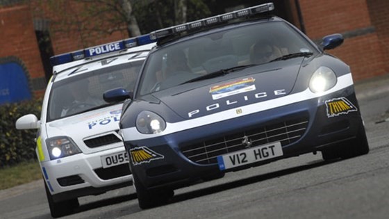 A dark blue Ferrari 612 Scaglietti followed by a white UK police car.