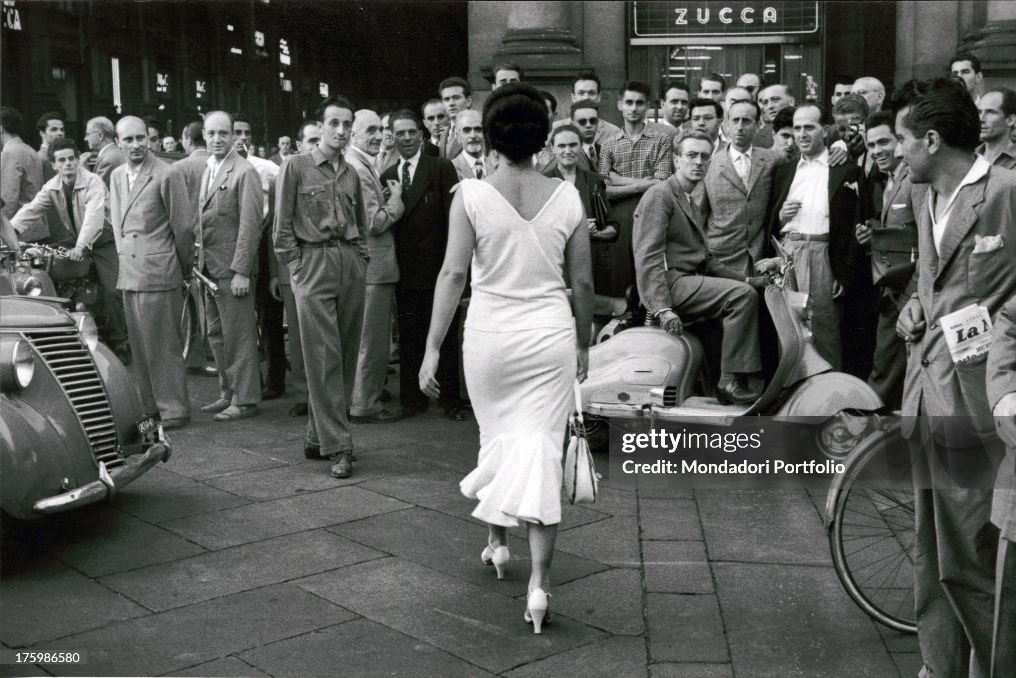 Young actress and circus performer Moira Orfei walks towards the Galleria Vittorio Emanuele II in Milan in 1954 while a large group of men turn to look at her.