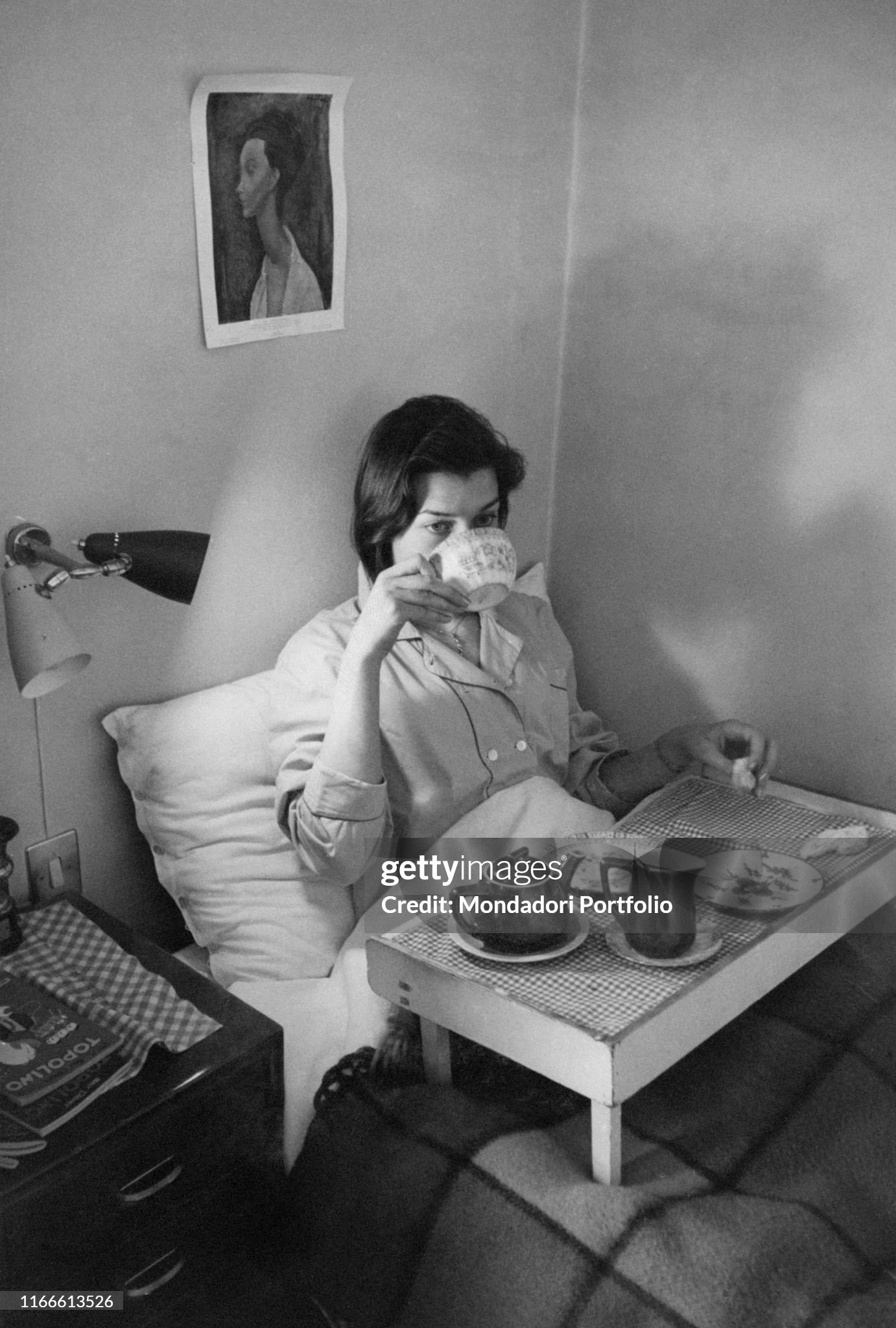A girl in pajamas has breakfast in her bed, on her legs a tray, in Milan in 1956; the photographer portrays her while she is raising a cup up to her lips. 