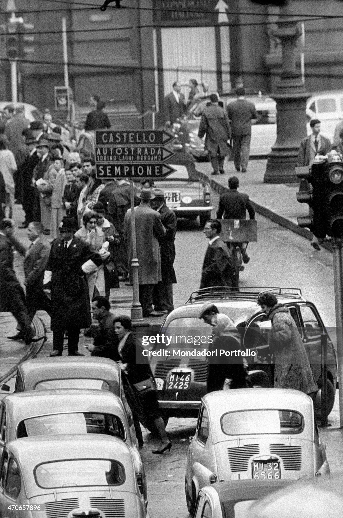 View of a street crowded with passersby waiting for the tram to arrive and filled with cars in Milan, Italy, in 1960; in the middle, a traffic sign gives information about the directions to follow to reach Castello, Sempione and the motorway. 