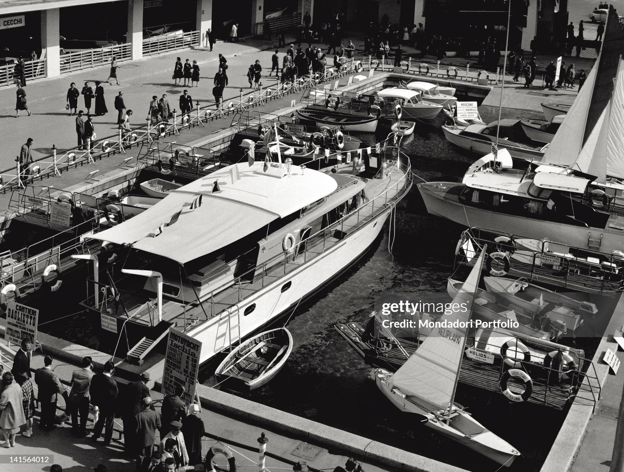 By the motor-boating sector of the 38th edition of Milan Trade Fair, a crowd of visitors is watching a big yacht moored next to motor-boats and sailing boats in 1960. 