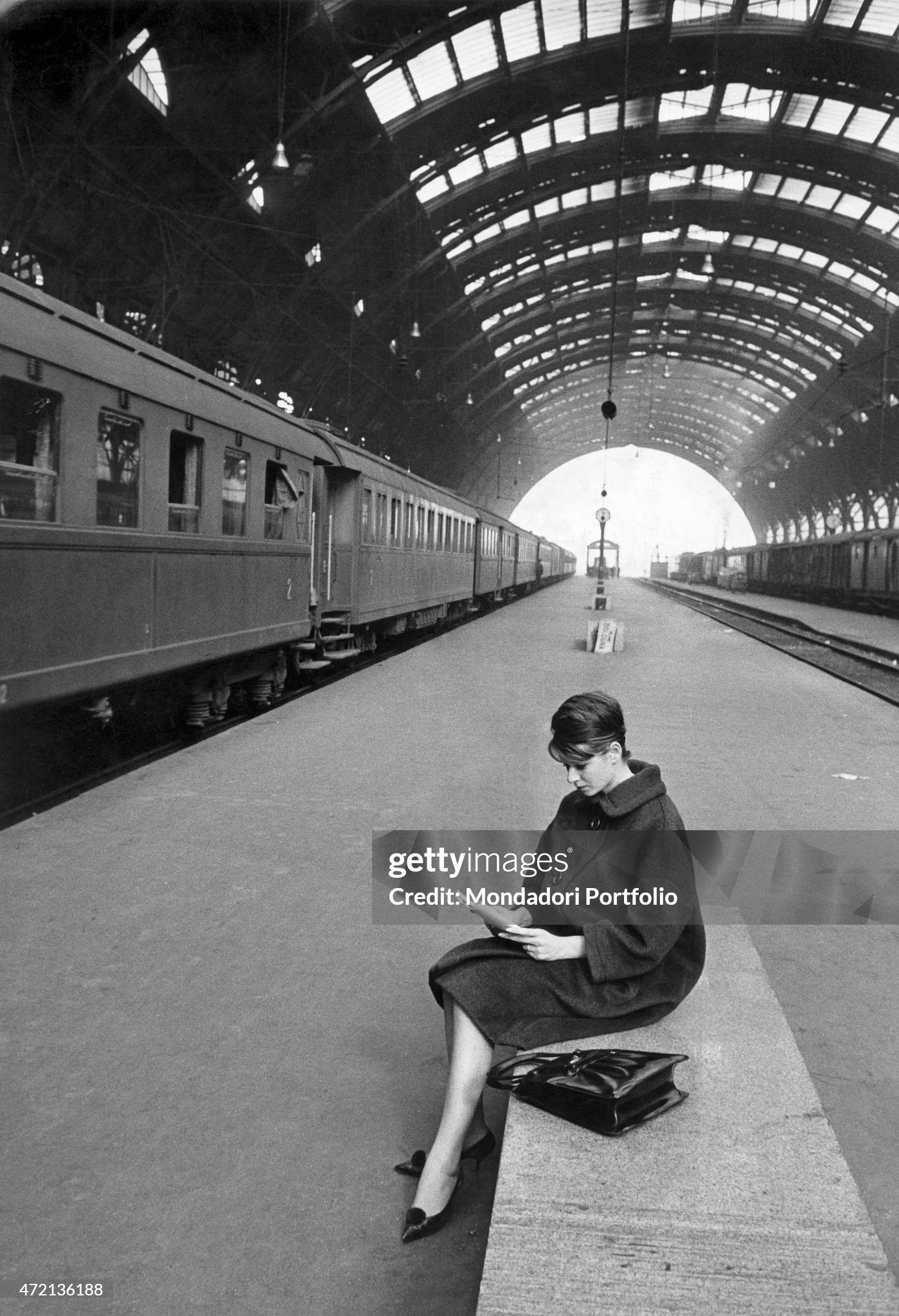 A young woman waiting sitting on a bench at Milano Centrale railway station in 1961. 