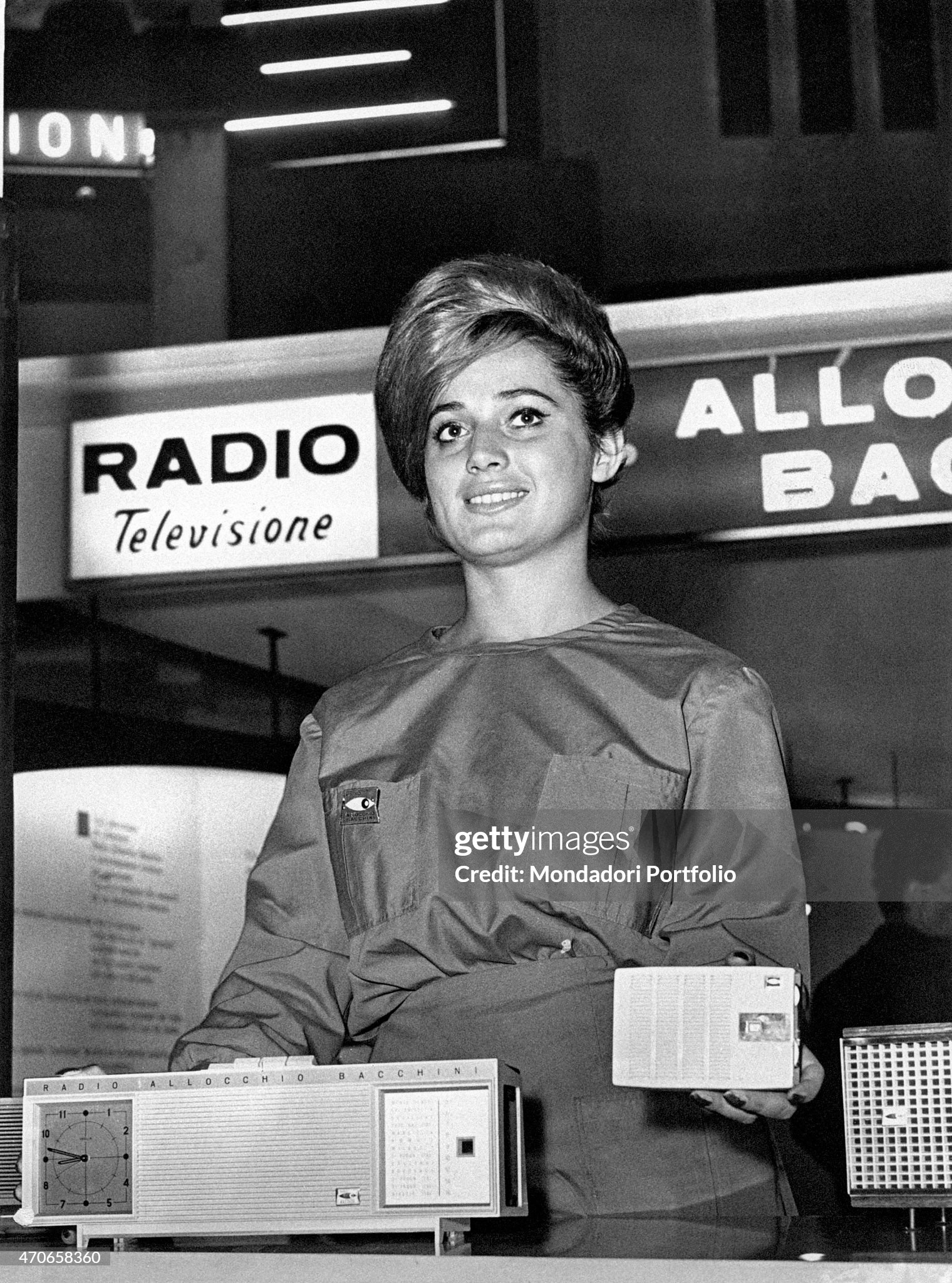 In the occasion of an exposition of radios, television and electrical appliances, a smiling hostess shows Cucciolo, a seven transistors radio that adopts the functions of an alarm clock and a timer connecting them to a radio station alterable according to the client's favourite music; assembled by the Allocchio Bellini and sold at 21.000 liras, it also can be set in the dashboards. Milan, Italy, October 1961. 
