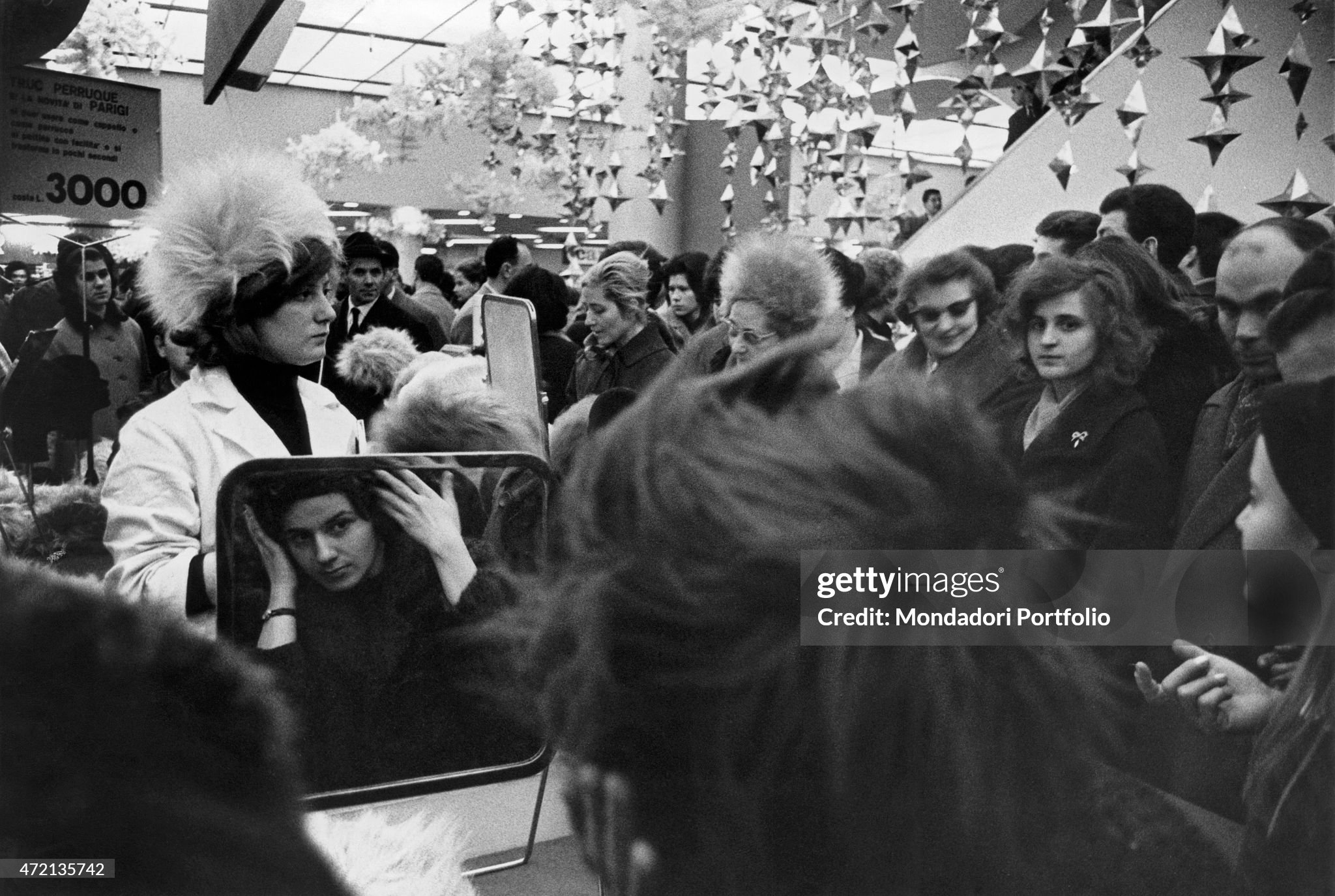 A young woman trying on a hat at La Rinascente, crowded during Christmas shopping time, in Milan on 01 December 1962. 
