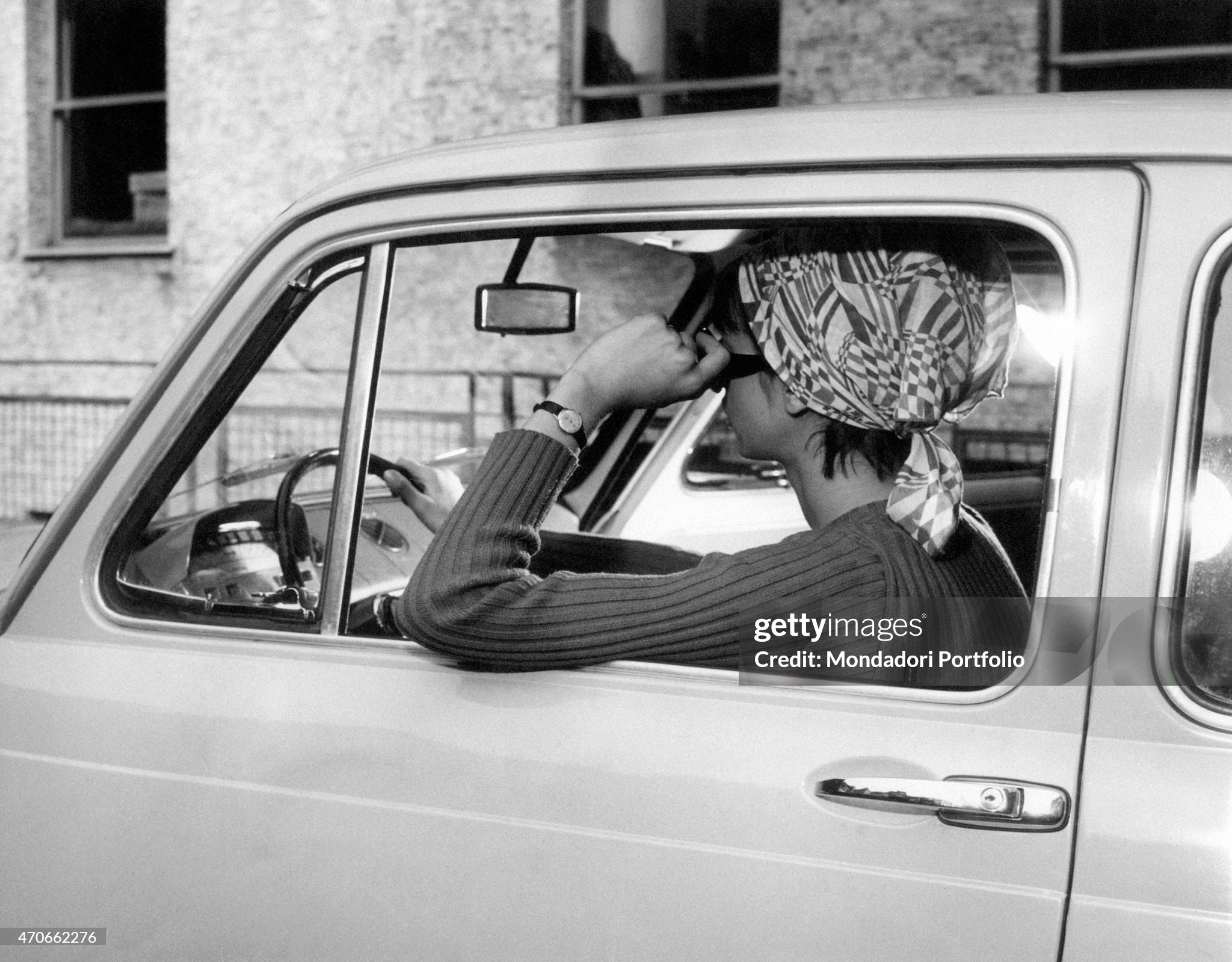 A thoughtful girl with a foulard in her hair and sunglasses drives her city runabout, leaning her arm out of the open window car in Milan on 20 April 1966. 