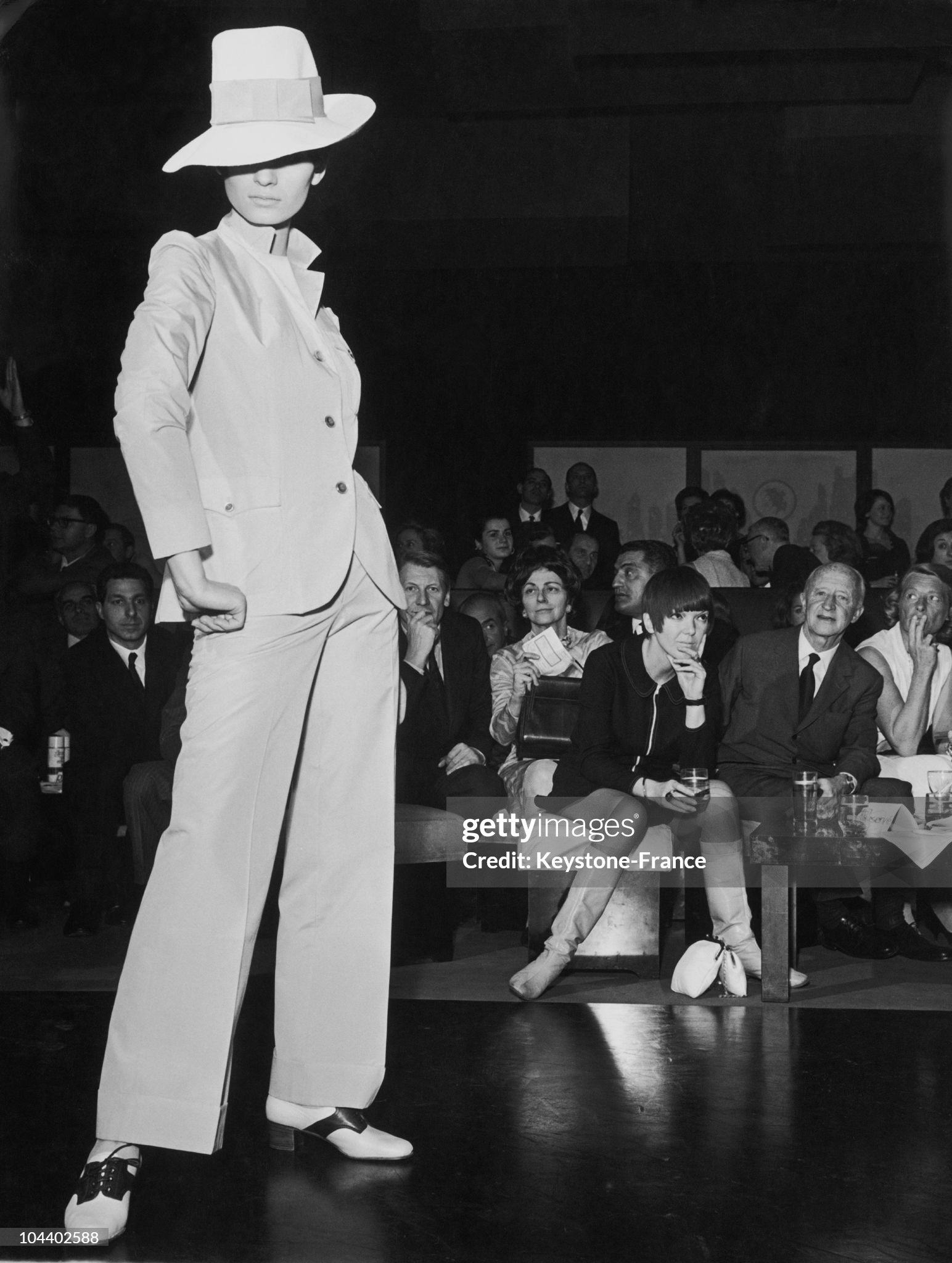 Mary Quant (seated in the background) is presenting a lady's suit from her collection Viva Viva during Milan fashion show in 1967. This suit is composed of a white jacket and baggy pants. The model is also wearing a large boards hat and bicolored shoes with a light heel. 