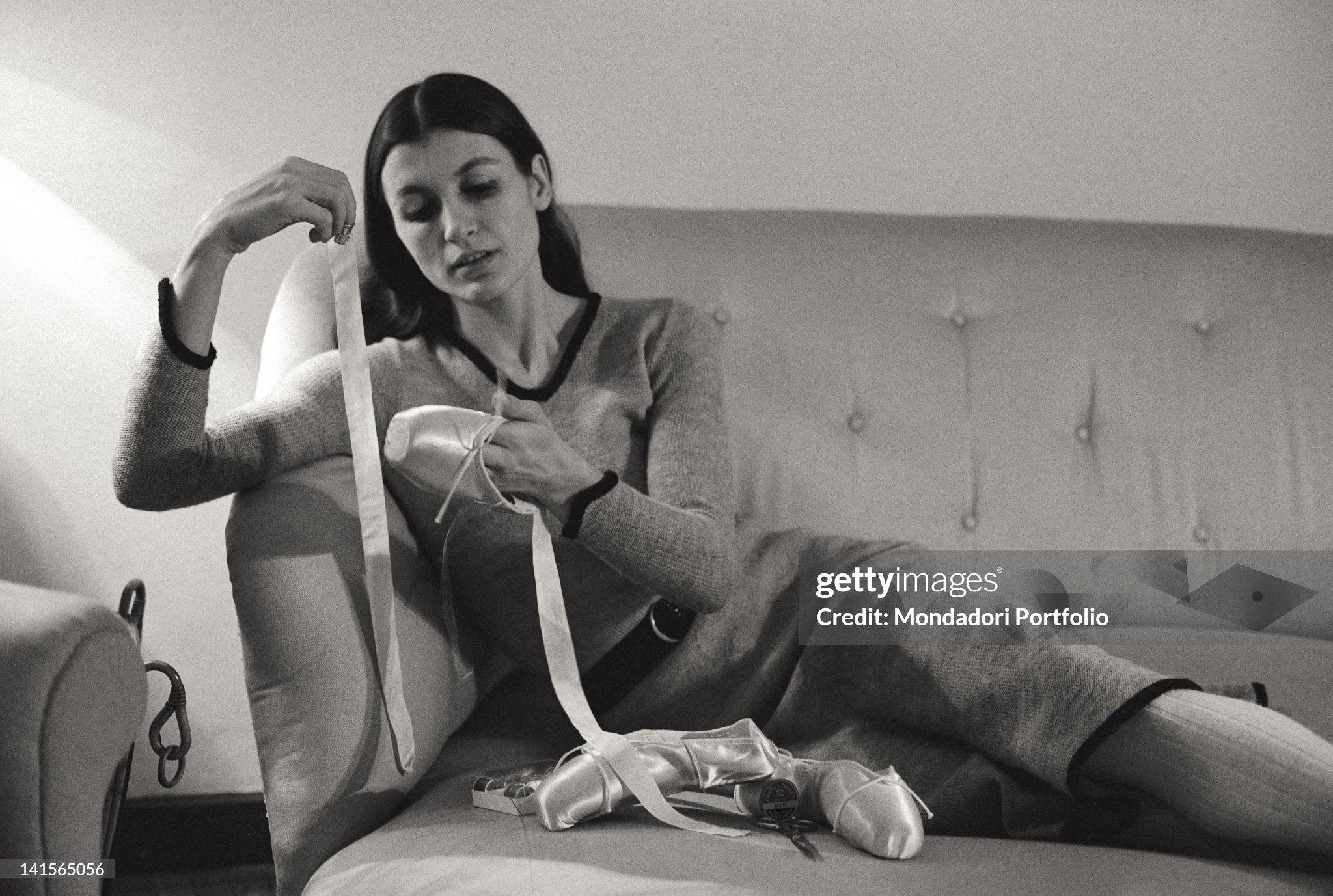 Portrait of the ballet dancer Carla Fracci, sat down on the sofa of her mansard while she is sewing by hand, applying some silk ribbons on her dancing shoes. Milan, February 1969. 