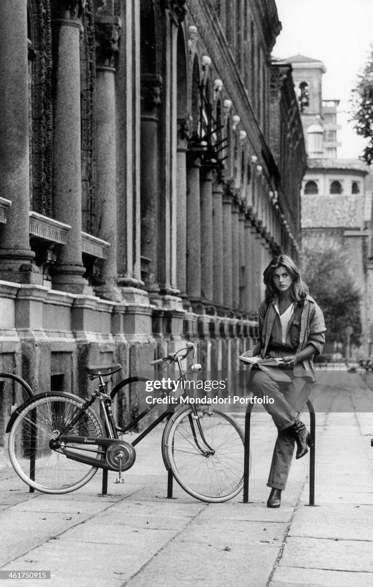 A girl standing beside her bicycle holding an open book outside the University of Milan in 1970s. 