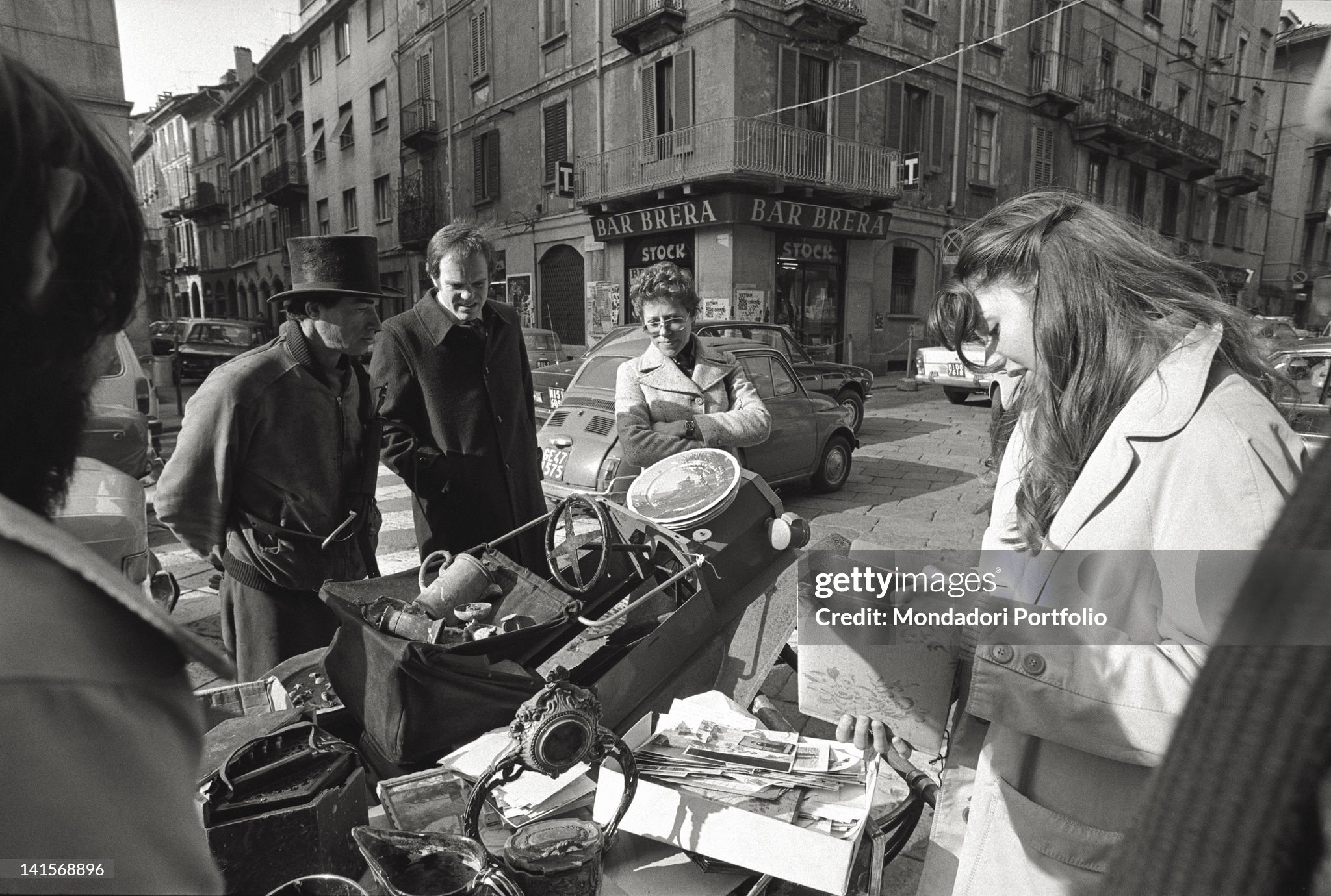 People buying from antiques barrows in 1974; interesting local characters. In the background the Brera bar bookshop in one of the principal streets of Brera, the Latin Quarter of Milan, with its luxury restaurants and sophisticated bars and discotheques which have replaced the picturesque 'low-life' cafes of the artists of the bohemian and hippy period. 