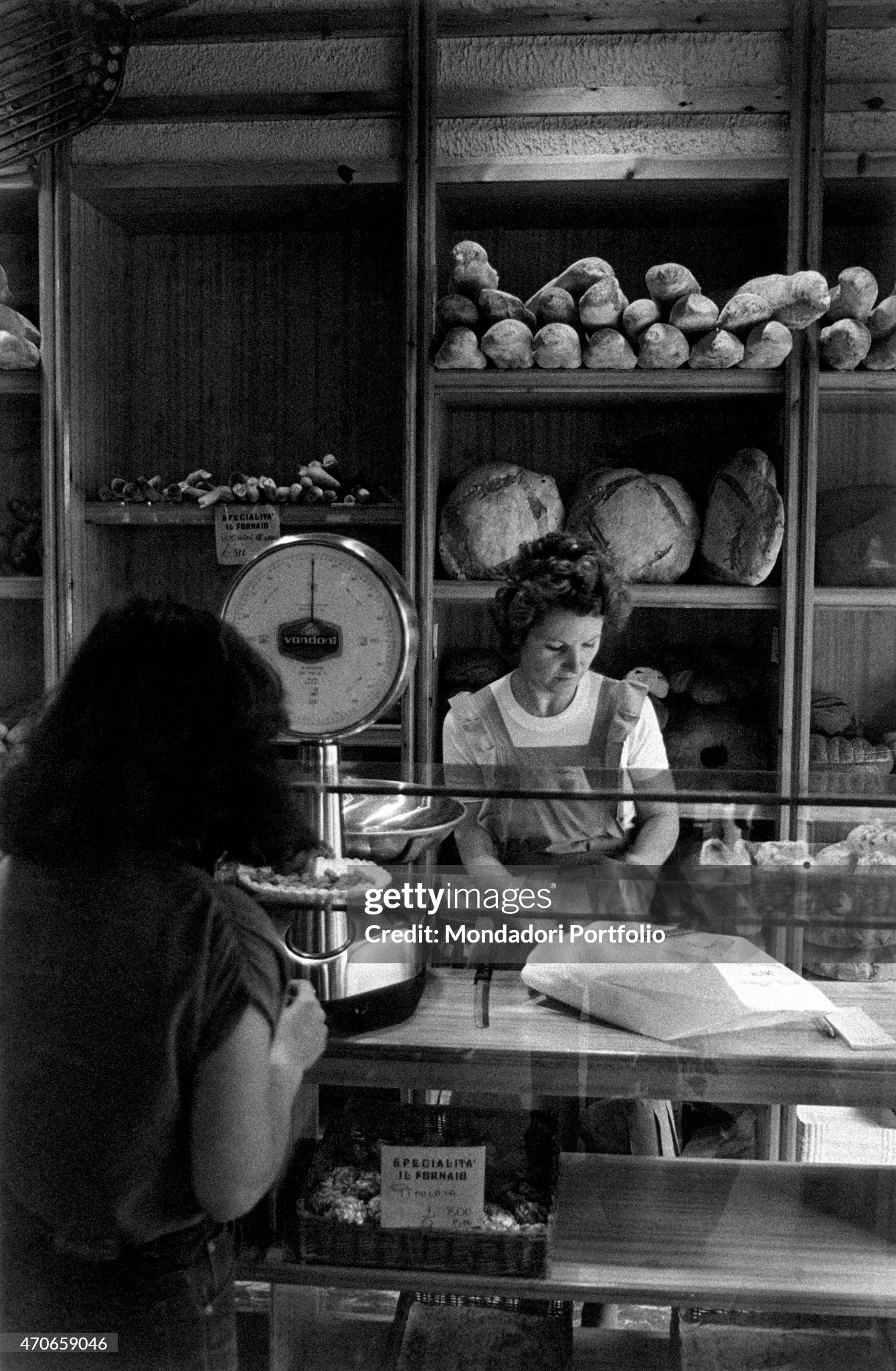 In a bakery the clerk puts some bread in a small bag while the customer, standing in front of her, waits. Milan, Italy, 1980. 