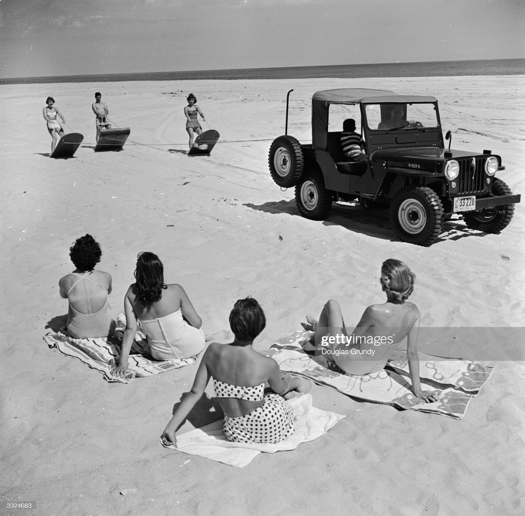 Circa 1955: an all-terrain vehicle towing three girls on sand skis across a beach on the east coast of America.