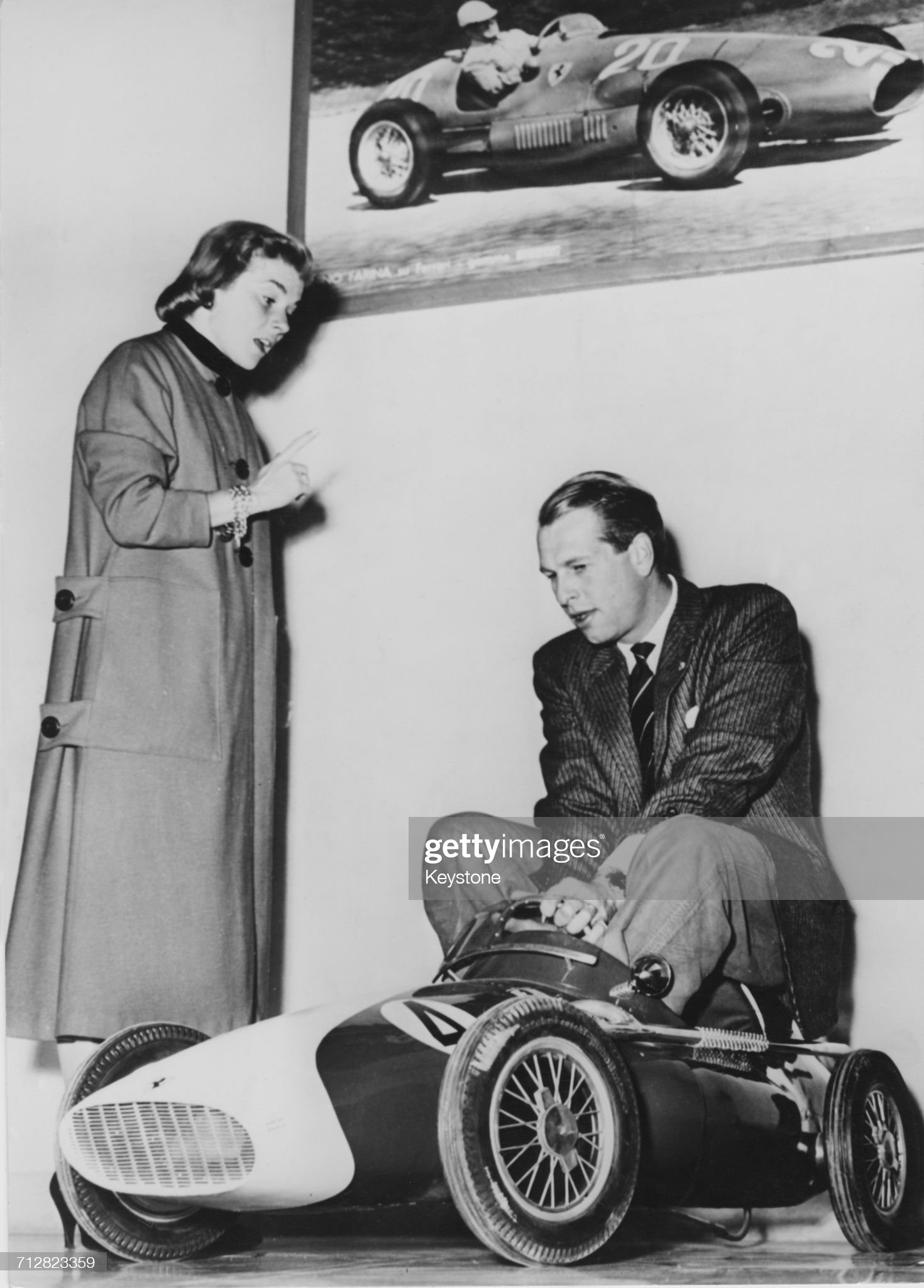 Peter Collins, team driver for the Scuderia Ferrari, plays on a toy replica Ferrari as his wife Louise King watches.