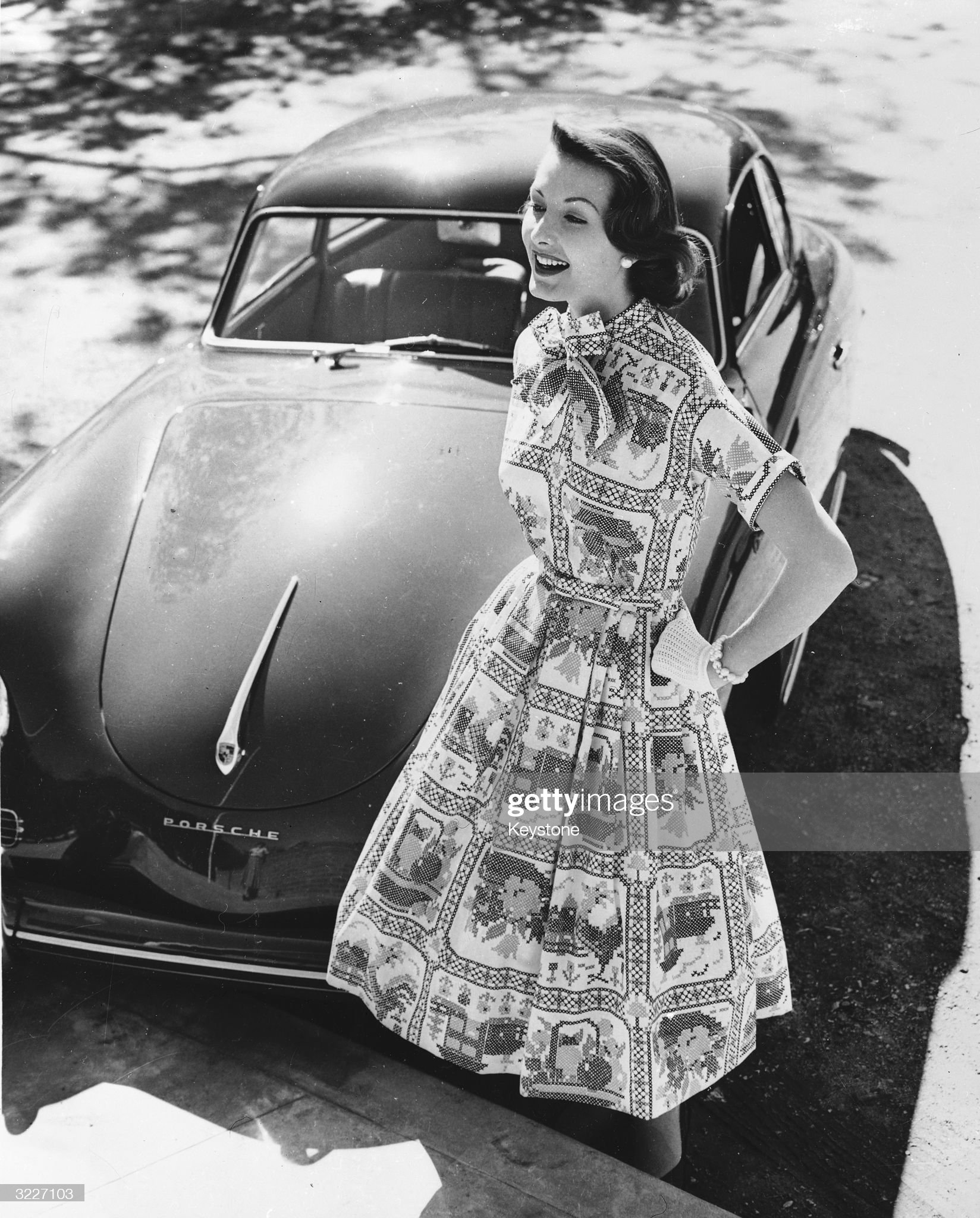 A woman models a cotton printed dress next to a Porsche sportscar in 1957.