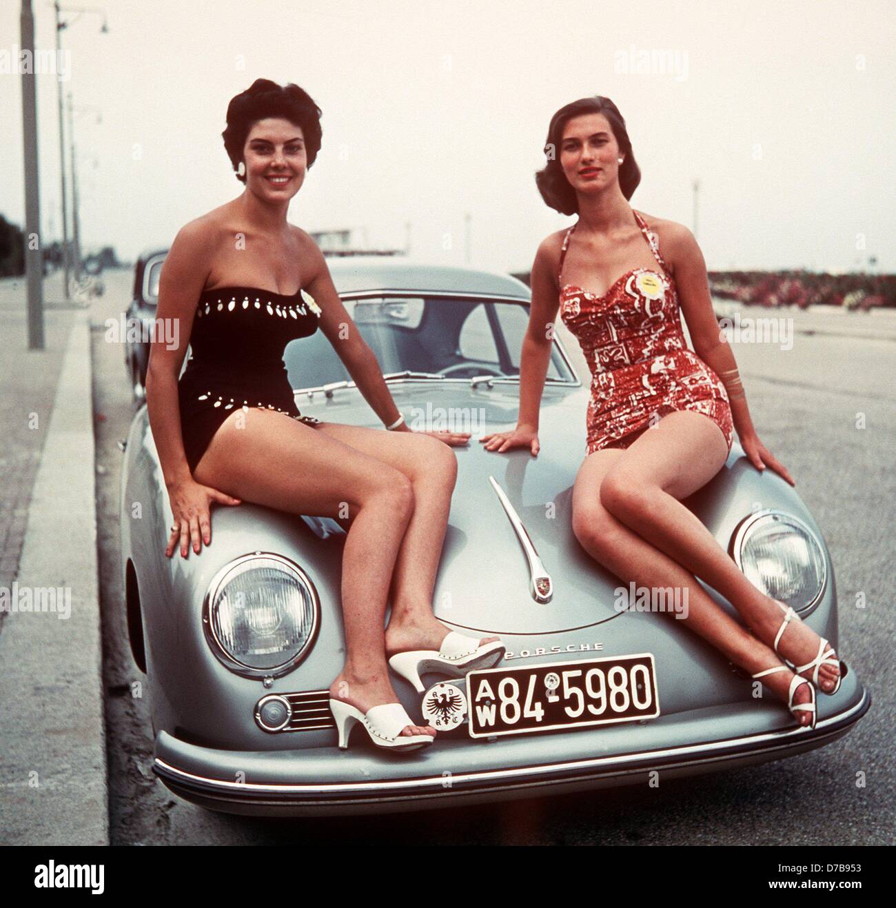 Two young ladies wearing a swim suit sit on the bonnet of a Porsche 356.
