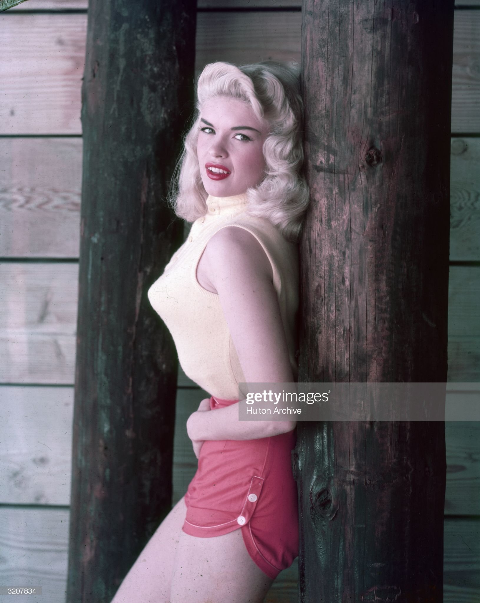 Circa 1955: portrait of American actor Jayne Mansfield, wearing a sleeveless sweater, pointy bra and red shorts, leaning against a wooden post, 1950s.