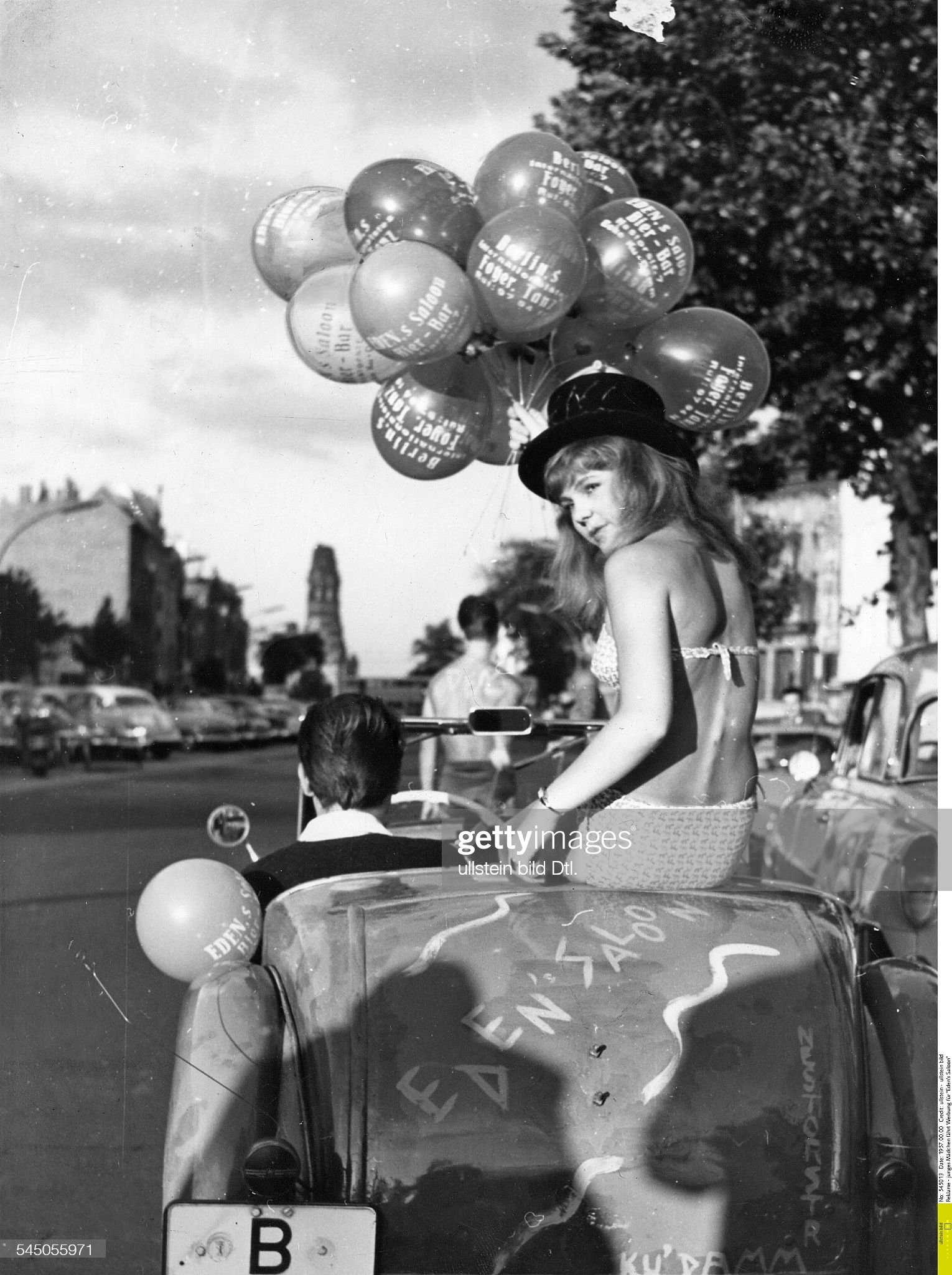A young girl in a bikini holds balloons in her hand and sits in a cabriolet driving on Kurfürstendamm in Berlin, advertising 'Eden's Saloon' in 1957. 