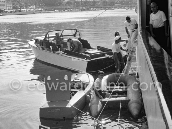 In the Monaco harbor in 1957 a young Alexander Onassis in a striped shirt sits on the pontoon of an inflatable dinghy, one of the tenders for the yacht ‘Christina’. He looks up as crew members descend a ladder from the main vessel's high hull. Moored alongside are other support boats, including a sleek motor launch with the name ‘CHRISTINA O.A.’ visible on its side. 