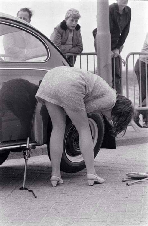A vintage woman changes the tire on a Volkswagen Beetle.