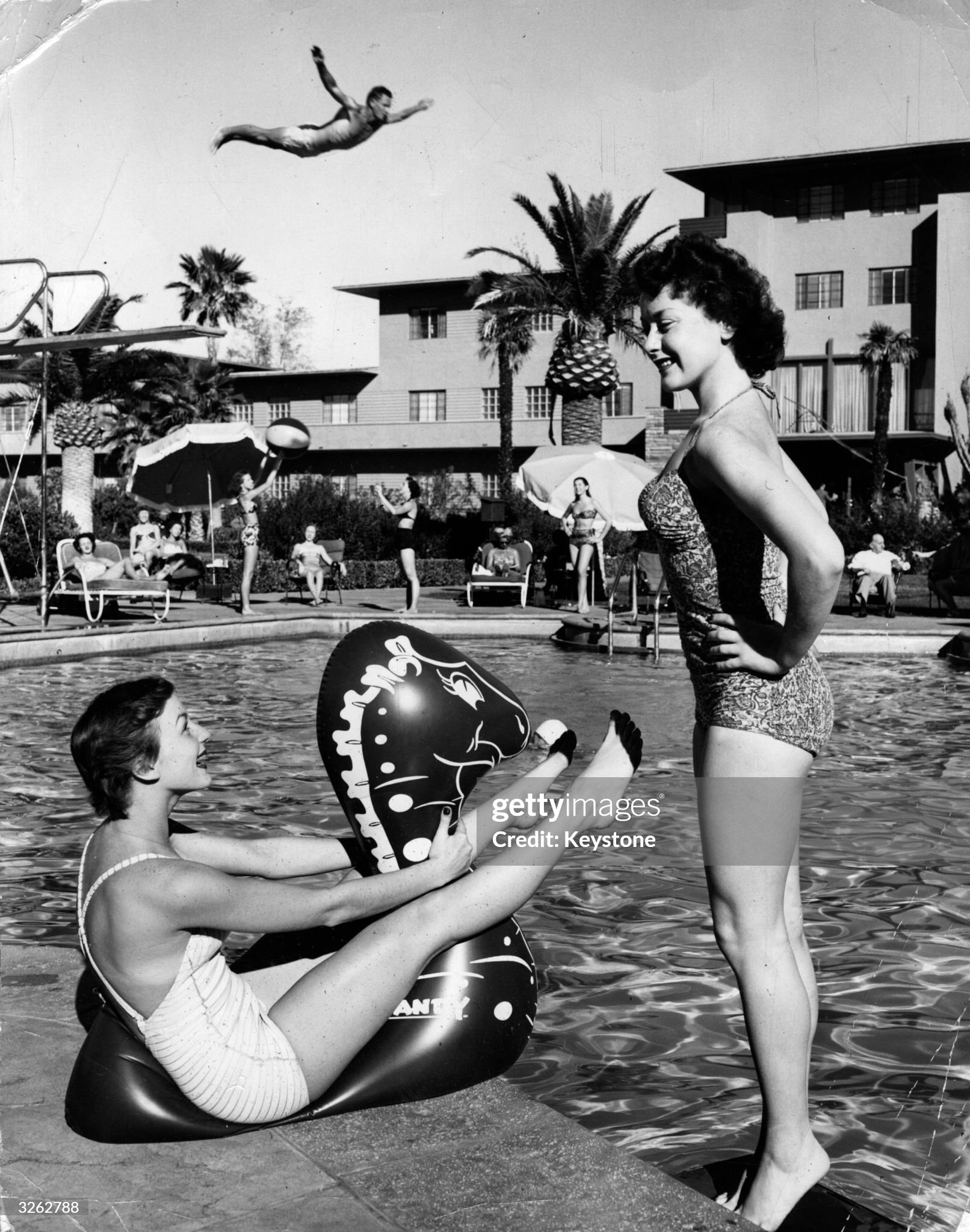 Two young women enjoying themselves by the pool at a Las Vegas holiday resort, while a man performs a spectacularly high dive in the background, circa 1955. 