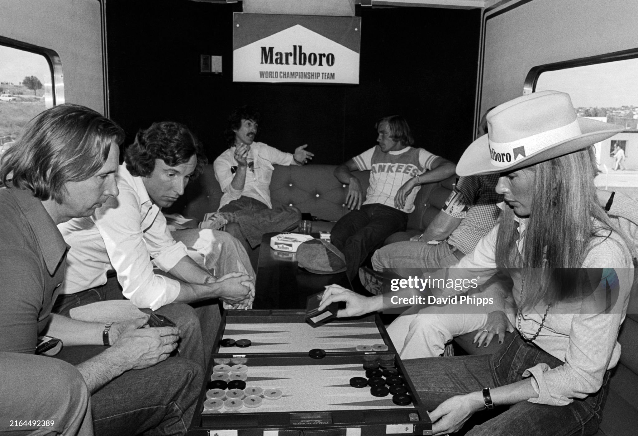 George Harrison enjoys a cigarette and a chat with race retiree James Hunt, McLaren, in the Marlboro motorhome as Nina Rindt plays backgammon, at the Spanish Grand Prix in Jarama on 08 May 1977. 