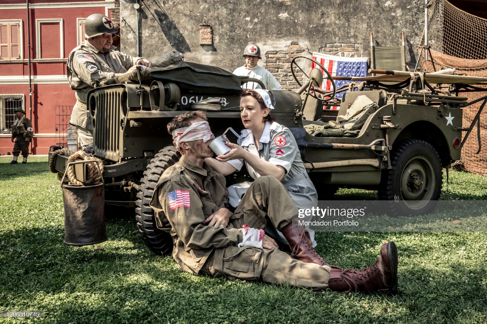 Young soldier shot in the face resting against the wheel of a Willis Jeep and being helped with drinking by a nurse.