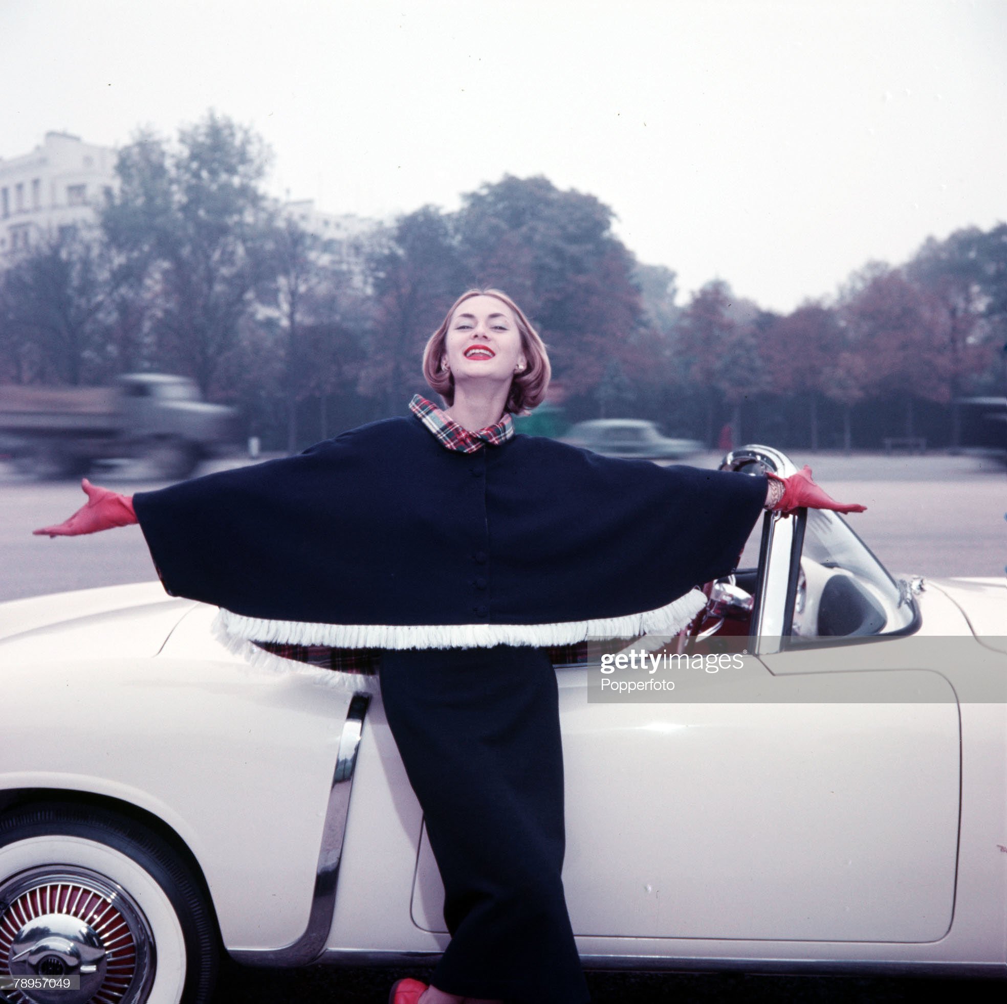 An elegantly dressed woman is pictured wearing a two piece blue outfit and cape as she stands next to her open-topped sports car, circa 1960.
